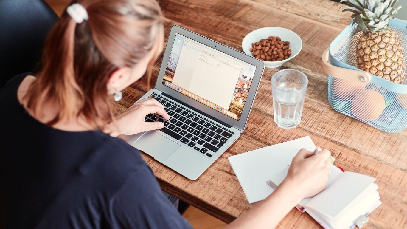 Femme travaillant sur un ordinateur portable, prenant des notes à une table avec un verre d'eau, des amandes et des fruits – préparation au traitement FIV pour femme célibataire