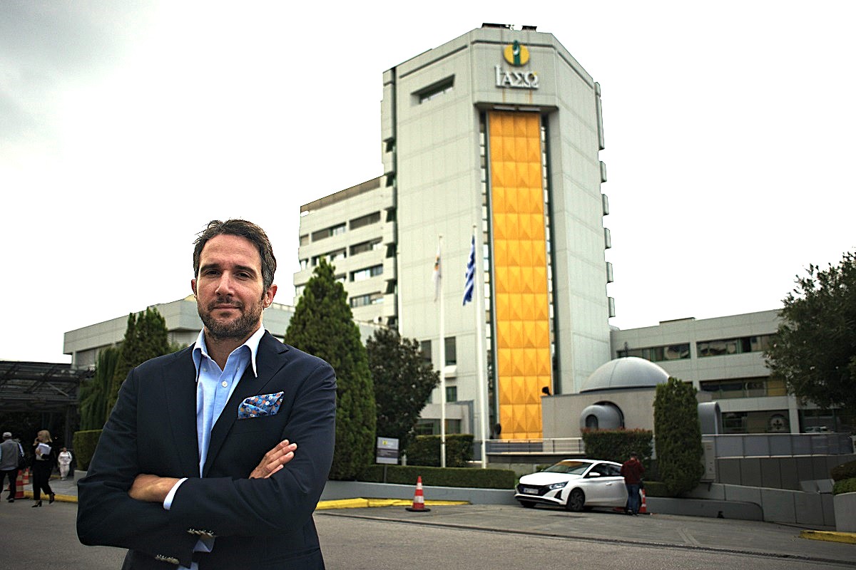 Dr Panagiotis Drakopoulos devant l'hôpital IASO d'Athènes : homme en costume-cravate, drapeau grec, bâtiment moderne à la façade jaune.