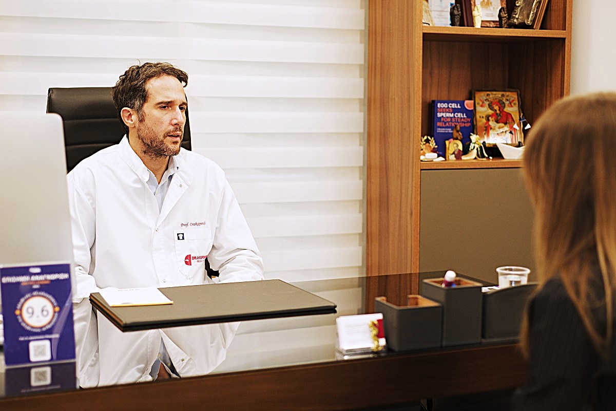 Le Dr Panagiotis Drakopoulos conseille une patiente en médecine de la reproduction : médecin en blouse blanche à son bureau, femme aux cheveux longs, étagère de livres en arrière-plan